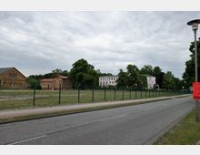 Straßenansicht mit einem Gehweg und einer Fahrbahn im Vordergrund, dahinter eine umzäunte Wiese. Im Hintergrund stehen mehrere Gebäude, darunter ein größeres weißes Haus und braune Gebäude mit roten Dächern, umgeben von Bäumen.