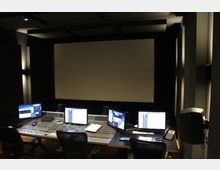 Tonstudio mit einer großen Leinwand im Hintergrund, einem Mischpult und mehreren Monitoren auf zwei Arbeitsplätzen im Vordergrund. Der Raum ist schalldicht gestaltet und schwach beleuchtet, mit modernen Wandpaneelen und integrierter Beleuchtung.