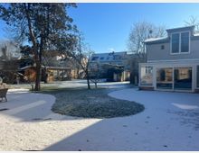 Verschneiter Garten mit Rasen- und Terrassenflächen, umgeben von Gebäuden, darunter ein modernes Wohnhaus mit großen Fenstern und ein Holzschuppen. Im Hintergrund sind weitere Wohnhäuser und kahle Bäume zu sehen.