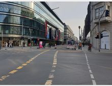 Straßenkreuzung in einer urbanen Umgebung mit großen modernen Gebäuden, darunter ein Einkaufszentrum mit gläserner Fassade auf der linken Seite. Verkehrsmarkierungen, Fußgänger, Radfahrer und Baustellenabsperrungen prägen die Szene.