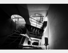 Schwarz-weiß-Fotografie eines Treppenhauses mit mehreren Metall- und Betontreppen, geschwungenen Wänden und bogenförmigen Durchgängen. Oberhalb sind Geländer und Gitter sichtbar, die den Raum strukturieren.