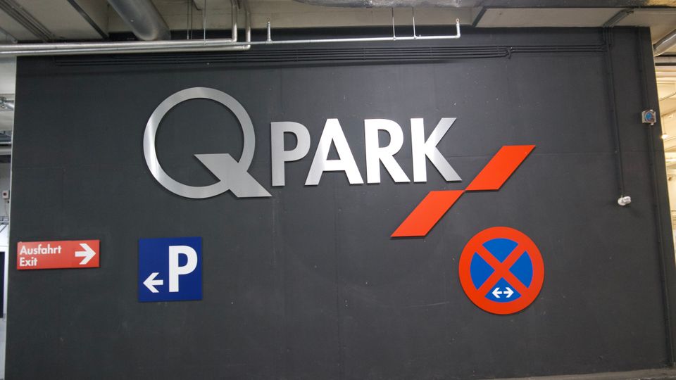 Eine Wand in einem Parkhaus mit dem Schriftzug „Q-PARK“ in silbernen Buchstaben, ergänzt durch ein rotes Logo. Darunter befinden sich ein Parkschild mit einem blauen „P“ und Pfeil nach links sowie ein rot-blaues Halteverbotszeichen. Ein Schild mit der Aufschrift „Ausfahrt / Exit“ zeigt nach rechts.