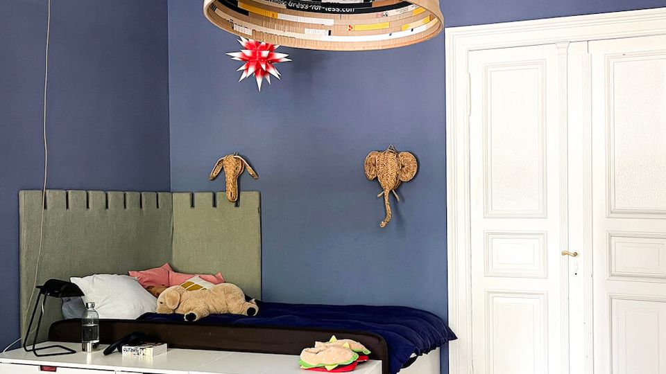 Bedroom interior with deep blue walls, ornate white ceiling molding, and a large cylindrical pendant light hanging over a bed with an upholstered headboard. A low white drawer unit sits at the foot of the bed, with white paneled double doors and a herringbone wood floor completing the room.