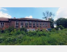 Backsteinbau mit zerstörten Fenstern und einem stark beschädigten Dach, umgeben von dichter Vegetation und Wildwuchs; Graffiti bedeckt Teile der Fassade, und im Hintergrund sind Bäume unter einem bewölkten Himmel sichtbar.