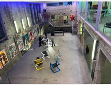 Industriehalle mit Kunstgalerie: Ein großer Raum mit freiliegenden Backsteinwänden, an denen bunte Kunstwerke hängen. Moderne Metallmöbel und eine gläserne Galerieebene prägen das Design, während neonartige Beleuchtung und ein Betonboden eine industrielle Atmosphäre schaffen.