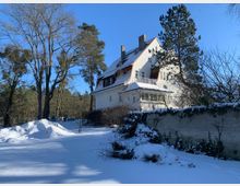Schnee bedecktes Grundstück mit einem großen weißen Wohnhaus im Landhausstil, umgeben von hohen Bäumen und einer weiß verputzten Gartenmauer. Der Himmel ist klar und blau, und der Garten ist komplett mit Schnee bedeckt.