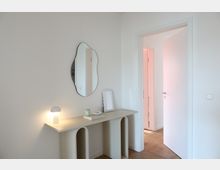 Minimalistisches Interieur mit einer beigefarbenen Konsole, auf der eine kleine Lampe, ein Kerzenhalter und ein dekorativer Gegenstand stehen. Darüber hängt ein organisch geformter Spiegel, während rechts eine geöffnete Tür zu einem angrenzenden Raum führt.