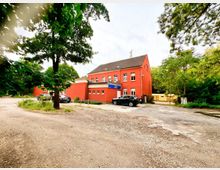 Zweigeschossiges, rot gestrichenes Gebäude mit Satteldach und mehreren Fenstern, davor ein kleiner Parkplatz mit Zufahrt. Das Haus liegt in einer grünen Umgebung mit großen Bäumen und dichtem Bewuchs am Rand des Geländes.