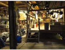 Innenansicht einer rustikalen Scheune oder Werkstatt mit Holzboden, Regalen und Hängeseilen. Verschiedene Werkzeuge, Bootelemente und Ausrüstungsgegenstände sind im Raum verteilt, beleuchtet durch warmes Licht.