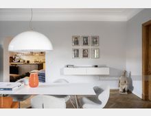 Modernes Esszimmer mit hellgrauen Wänden, einem weißen Tisch, gepolsterten Stühlen und einer großen, halbkugelförmigen weißen Hängelampe. An der Wand hängen sechs gerahmte Porträtbilder, darunter ein schwebendes Sideboard, und rechts steht eine kleine Terrakotta-Kriegerstatue. Im Hintergrund ist eine offene Küche sichtbar.