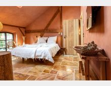 Gemütliches Schlafzimmer mit warmen Terrakotta-Wänden, offenem Holzbalken an der Decke und einem Bett mit weißer Bettwäsche. Der Raum verfügt über eine Holztür, rustikalen Steinboden, ein Fenster mit Blick nach draußen und Holzmöbel, darunter ein Tisch mit einer dekorativen Schale.