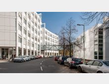 Moderne Bürogebäude mit weißer Fassade und großen Fenstern, verbunden durch eine gläserne Fußgängerbrücke. Davor eine Straße mit geparkten Autos und einigen kahlen Bäumen.