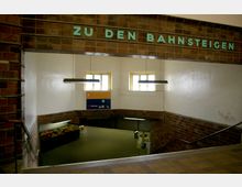 Eingangsbereich zu einem Bahnhofsuntergeschoss mit braunen Kachelwänden und dem Schriftzug „ZU DEN BAHNSTEIGEN“. Zwei rechteckige Fenster und Hängelampen erhellen den Raum, während im unteren Bereich Menschen in der Nähe eines Fahrkartenautomaten stehen.