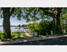 Blick auf eine kleine Bootsanlegestelle am Ufer eines Sees, umgeben von dichter Vegetation und schattenspendenden Bäumen. Im Hintergrund ist ein weißes Ausflugsschiff auf dem Wasser zu sehen, während ein Weg im Vordergrund durch Geländer gesichert ist.