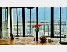 Ein heller Raum mit einem Holztisch im Vordergrund, darauf eine Vase mit roten Blumen, eine Obstschale und ein Laptop. Durch große Glasfenster im Hintergrund sieht man eine Terrasse mit Möbeln und im weiteren Blick den Berliner Fernsehturm.