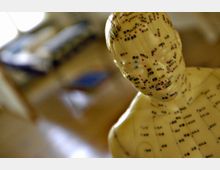 Nahaufnahme einer Akupunktur-Modellfigur, die mit Punkten und chinesischen Schriftzeichen zur Darstellung von Akupunkturpunkten markiert ist. Im unscharfen Hintergrund ist ein heller Raum mit einem Bett und einem kleinen Tisch zu erkennen.