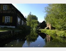 Ein schmaler Kanal mit klarem Wasser, flankiert von einem Fachwerkhaus mit gelben Fensterläden auf der linken Seite und einer Backsteinscheune sowie Bäumen auf der rechten Seite, unter blauem Himmel.