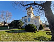 Hellgelbes, historisch wirkendes Gebäude mit Säulen und einem Turm, umgeben von gepflegtem Rasen, Bäumen und Büschen. Im Vordergrund stehen Parkbänke, während der blaue Himmel die Szene beleuchtet.