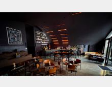 Eine moderne Bar mit dunkler Beleuchtung und stilvoller Einrichtung. Der Raum hat eine gewölbte Decke, eine beleuchtete Regalwand mit Flaschen hinter der Theke, Tische und Stühle im Vordergrund sowie gemütliche Sitzgelegenheiten auf der rechten Seite.