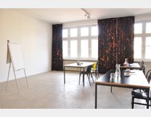 Ein heller Seminarraum mit weißen Wänden und großen Fenstern, die durch schwarze Vorhänge mit orangefarbenem Muster teilweise verdeckt sind. Im Raum stehen mehrere Tische mit Stühlen sowie ein Flipchart.