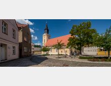 Stadtplatz mit einer historischen Kirche im Zentrum, gekennzeichnet durch ein gelbes Gebäude mit rotem Dach und einem hohen Kirchturm mit Uhr. Umgeben von gepflasterten Gehwegen, Bäumen und angrenzenden Wohnhäusern.