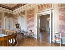 Prunkvoller Innenraum mit klassizistischer Gestaltung, verziert mit gemalten Wandreliefs, einer Holzbalustrade, Marmordekor und aufwendigen Stuckarbeiten. Ein Porträt auf einer Staffelei sowie elegante Stühle und ein Kronleuchter im angrenzenden Raum runden das historische Ambiente ab.