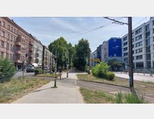 Straßenansicht in einem städtischen Viertel mit mehrstöckigen Wohn- und Geschäftsgebäuden auf beiden Seiten, Gehwegen, Straßenbahngleisen und einer Ampel. Im Hintergrund sind Bäume und eine lichte Allee sichtbar, daneben eine Werbetafel an einem modernen Gebäude.