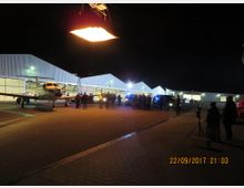 Nachtszene auf einem Flugplatz mit mehreren Hangars im Hintergrund, beleuchtet von künstlichem Licht. Im Vordergrund steht ein kleines Flugzeug und eine Gruppe von Menschen neben Fahrzeugen, während Kamera-Equipment am Rand positioniert ist.