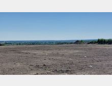 Weites, offenes Brachland mit kahler Erdoberfläche und vereinzeltem Müll; im Hintergrund eine von Bäumen bewachsene Hügellandschaft und ein klarer blauer Himmel.