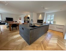 Moderne Wohnküche mit großem Küchenblock aus dunklem Stein, beigefarbenen Einbauschränken und einem Fenster mit Blick nach draußen. Der Raum hat hellen Fischgrätparkettboden, einen Esstisch mit Stühlen im Hintergrund sowie eine offene Verbindung zum Wohnbereich mit Sofa und Fernseher.