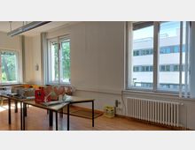 Innenraum eines einfachen Büros mit Holzboden, mehreren Fenstern und einem Heizkörper unterhalb der Fenster. Auf Tischen stehen diverse Kartons, Verpackungen und Gegenstände. Durch die Fenster ist ein modernes, mehrstöckiges Gebäude und Bäume zu sehen.