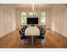 Konferenzraum mit einem langen, hellen Tisch und zwölf schwarzen Stühlen. Im Hintergrund befinden sich große Fenster mit Blick auf grüne Bäume sowie ein großer Bildschirm auf einem Standfuß, der zwischen den Fenstern platziert ist.