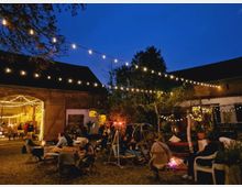 Ein stimmungsvoller Hof bei Nacht mit hängenden Lichterketten, rustikalen Backsteingebäuden und offenen Holztoren. Menschen sitzen auf Stühlen und Sofas in kleinen Gruppen, teilweise um Feuerstellen, umgeben von Bäumen und Beleuchtung.