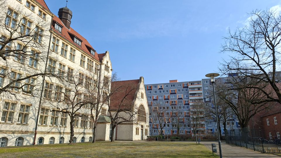 Innenhof mit Rasenfläche und Gehweg, umgeben von einem historischen hellen Gebäude mit steilem roten Ziegeldach, vielen Fenstern und einem kleinen Turm sowie einem hohen modernen Wohnblock im Hintergrund. Kahle Bäume, Laternen und Fahrradständer stehen am Rand des Platzes.