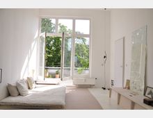 Helles Wohnzimmer mit einer großen, geöffneten Glastür, die zu einem Balkon mit Pflanzen führt. Der Raum ist minimalistisch eingerichtet mit einer niedrigen Liegefläche, Kissen, einem Spiegel an der Wand und hellen Wänden.