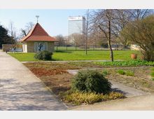 Kleiner Pavillon mit spitzem Ziegeldach und hellen Ziegelwänden in einer Parkanlage mit Rasenflächen, Bäumen und Gehwegen, im Hintergrund ein modernes mehrstöckiges Gebäude. Ein Fahrrad lehnt an einer Bank, und Büsche sowie Blumenbeete säumen die Wege.