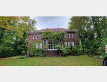 Backsteinhaus mit Satteldach inmitten eines Gartens, umgeben von Bäumen. Die Fassade ist mit Pflanzen bewachsen, und vor der Eingangstür führt eine breite Treppe nach oben.