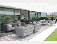 Moderne Terrasse mit grauen Polstermöbeln und einem rechteckigen Esstisch mit Stühlen unter einem hellen Sonnensegel. Im Hintergrund befinden sich große Glasfronten eines Hauses, die den Blick in den Innenbereich und auf einen Garten mit Rasen und Hecken freigeben.