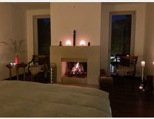 Gemütliches Schlafzimmer mit großem Bett im Vordergrund und offenem Kamin in einer schlichten, hellen Wandnische. Links und rechts stehen Sessel und kleine Tische mit Kerzen; hohe Fenster zeigen die dunkle Außenansicht.