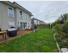 Blick in den Garten eines modernen Reihenhauses mit Holzterrasse, Esstisch und überdachten Gartenmöbeln. Daran schließt eine Rasenfläche mit Blumenbeeten, Sträuchern und Sichtschutzhecken an; im Hintergrund sind Nachbarhäuser und ein Spielgerät zu sehen.