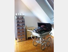 Ein Zimmer mit schräger Decke, hellen Wänden und Holzboden; links ein glänzendes, metallisches Möbelstück mit Schubladen, daneben eine kleine, beigefarbene Kommode. Im Vordergrund steht ein antiker Kinderwagen mit Büchern und Ordnern darin.