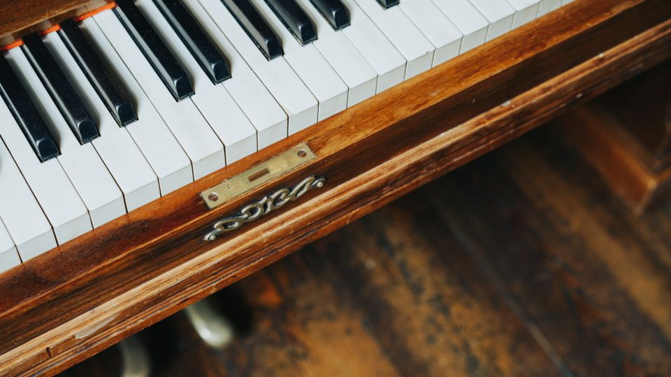Nahaufnahme der Tastatur eines Klaviers in einem Holzgehäuse, mit sichtbaren weißen und schwarzen Tasten sowie Metallbeschlag an der Front. Im Hintergrund ist ein abgenutzter Holzboden zu sehen.