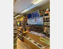 Ein kleiner Kiosk mit einem Verkaufsbereich, der aus einer Holztheke besteht. Darauf liegen Zeitungen und Zeitschriften, während im Hintergrund Zigarettenpackungen, Tabak und weitere Waren auf Regalen präsentiert werden. Über der Theke befinden sich Bildschirme mit Werbung, und eine Ecke zeigt ein Regal mit Süßigkeiten und kleinen Snacks.