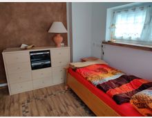 Ein schlichtes Schlafzimmer mit Holzboden, bestehend aus einem Einzelbett mit gemustertem Bettbezug in Rot- und Gelbtönen, einer Kommode mit Schubladen und integriertem Hi-Fi-System sowie einer Tischlampe. Rechts steht ein kleines Fenster mit weißem Vorhang, darunter eine Holzablage und eine goldene Leselampe.