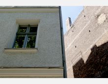 Ausschnitt eines Gebäudes mit einer hell verputzten Wand und einem rechteckigen Fenster, das grünes Laub reflektiert. Rechts im Bild befindet sich eine Ziegelwand mit kleinen Öffnungen und Schattenwurf.