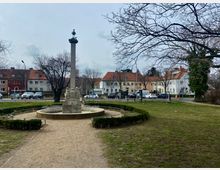 Ein kleiner Park mit einem zentralen Brunnen, bestehend aus einer Steinsäule mit einer Figur oben, umgeben von niedrigen Hecken. Im Hintergrund befinden sich mehrstöckige Wohnhäuser mit roten Ziegeldächern und geparkte Autos entlang einer Straße.