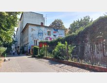 Zweistöckiges Gebäude mit einer bemalten Fassade, die bunte Graffiti und Illustrationen zeigt, darunter das Wort „CHIP“. Im Vordergrund verläuft ein Gehweg mit Pflastersteinen, flankiert von Grünflächen und einem bewachsenen Metallzaun. Im Hintergrund sind Bäume und weitere Gebäude zu sehen.