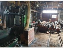 Holzwerkstatt mit schweren Maschinen, Laufbändern und gestapelten Baumstämmen auf Schienen. Im Hintergrund sind Holzbalkendecken, Werkzeug und große Fenster mit Lichteinfall sichtbar.