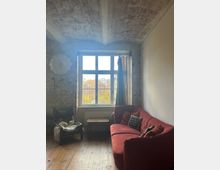 Wohnzimmer mit Backsteinwänden und gewölbter Decke. Im Vordergrund ein rotes Sofa, ein schwarzer Sessel mit Fellauflage und ein kleiner Holztisch, dahinter ein Fenster mit Blick auf bunte Herbstbäume.