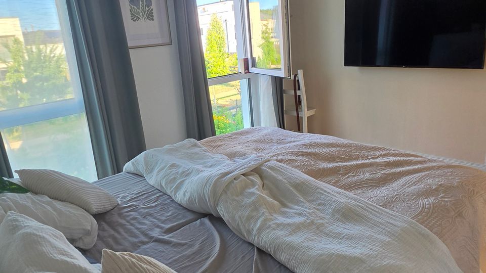Helles Schlafzimmer mit großem Bett und weißer Bettwäsche, grauen Vorhängen und bodentiefen Fenstern, von denen eines als Balkontür geöffnet ist. An der Wand hängt ein Flachbildfernseher, an der Decke eine geflochtene Pendelleuchte; draußen sind Grünflächen und Nachbargebäude sichtbar.